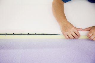 A technician sprays a sheet of pliable purple memory foam with a water-based adhesive and then carefully folds it over the other two foam layers and a steel spring grill to complete the backrest.