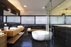 The sleek master bathroom features a freestanding tub, black mosaic tiles on the floor, and black natural stone on the walls. A natural oak vanity and oak accents add warmth to the otherwise cool and contemporary space, and a glass wall on the opposite side creates a sense of connection with the master bedroom. Photo 6 of Rishon Le Zion 3 modern home