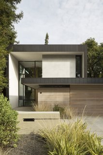 The slender two-story residence is clad in zinc, stucco, cedar, and glass. A low-lying garage sits at the front of the home.