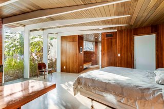 The master bedroom features floor-to-ceiling windows that soak the space in warm natural light. Thin wooden beams gradually run along the ceiling and extend to support a roof overhang.