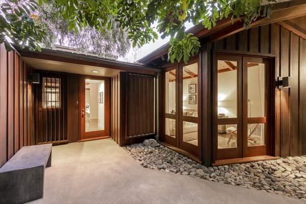 A view of the main entrance, which appears nestled within the trees. The minimalist exterior is clad in wood and surrounded by stones, creating a Zen-like retreat.