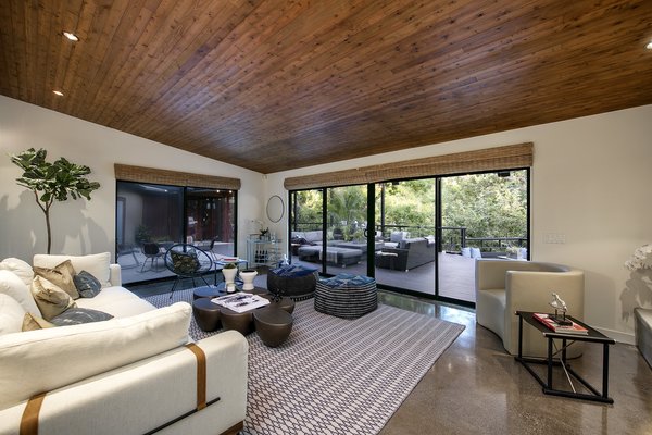 The large living area offers a striking wood-clad ceiling. Large sliding doors wrap around the room, providing access to the deck and framing views of the treetops.