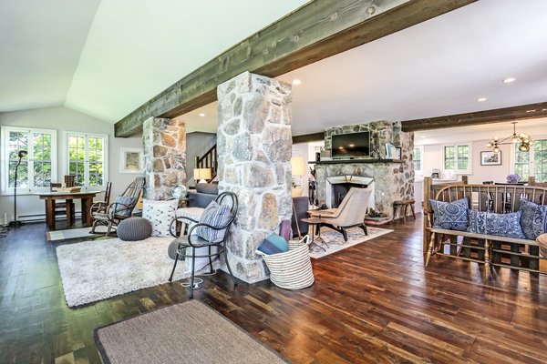 The sunroom flows into a large living room, which features a dramatic cut sone fireplace as the central focal point. Timber beams also run along the space, complementing the warm hardwood floors.