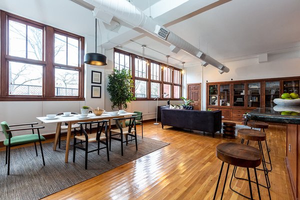 Newly restored hardwood floors and wooden trim run throughout the space, adding a warmth to the traditional industrial loft style. Bright upholstered chairs add a colorful touch, complementing the lush greenery throughout the space.