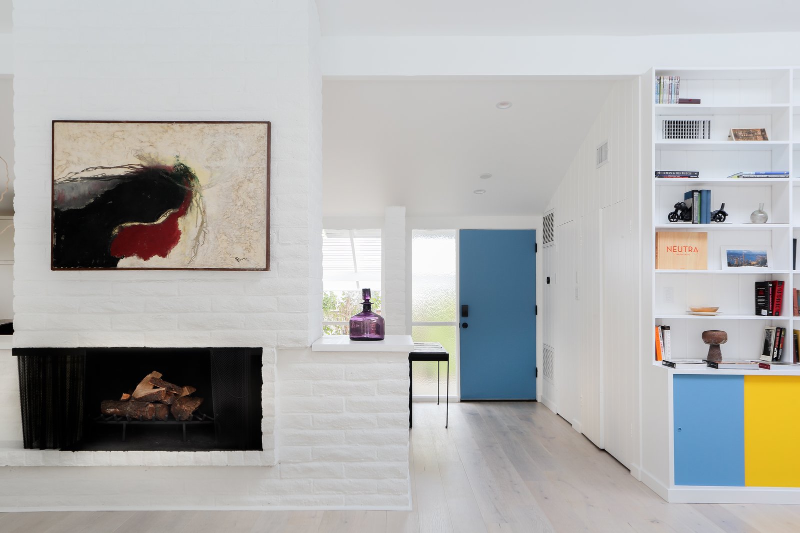 An original brick fireplace divides the living and dining room from the kitchen. Contrasting with an otherwise neutral color palette, pops of blue and yellow add flair to the built-in bookcase.
