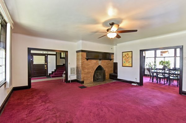 Much of the interior is currently hidden behind dated finishes and fixtures. Here, the living room features an original brick fireplace and wooden trim. Hardwood floors are also thought to be underneath the current carpeting.