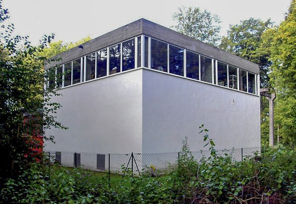 The smooth concrete facade is adorned by a series of ribbon windows along the top of three sides. Lush greenery surrounds the building and separates it from the main housing complex.