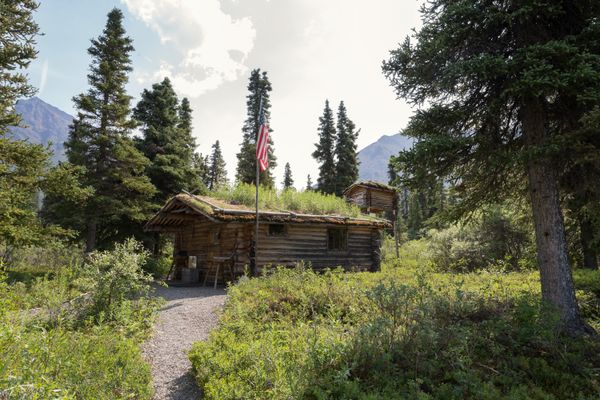 Dick Proenneke’s cabin and cache, surrounded by vegetation, which helps it blend into the forest. Proenneke used the cache to store goods.