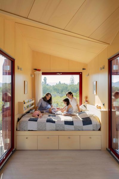 At one end of the plywood-paneled interior, a built-in bed holds four drawers—one for each member of the family. A picture window and two sets of glass sliders maximize the connection to the outdoors. The mattress is from Coco-mat, the lamp and clay pots are from Kave Home, the curtain is from London Fabric Company, the curtain pegs are from Kvadrat, and the prints are by Heath Robinson.