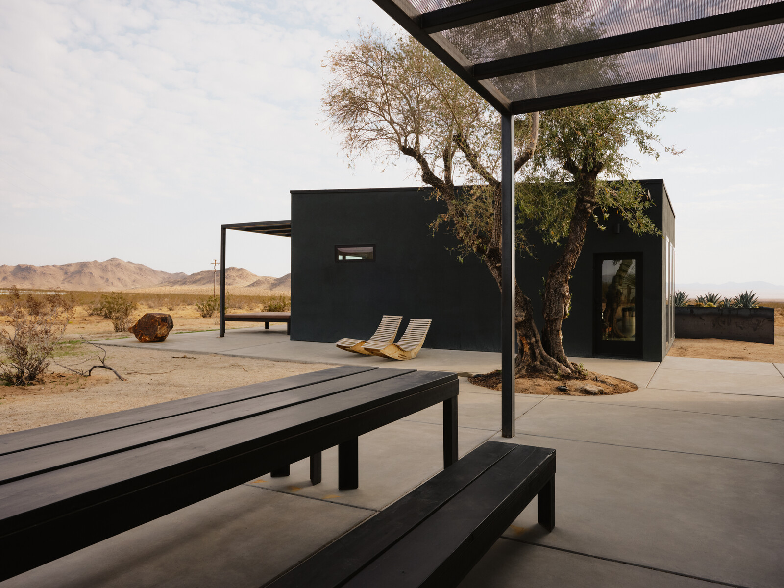 The main house and guest house are united by a shared patio. The table and benches in the foreground are made of steel and wood.