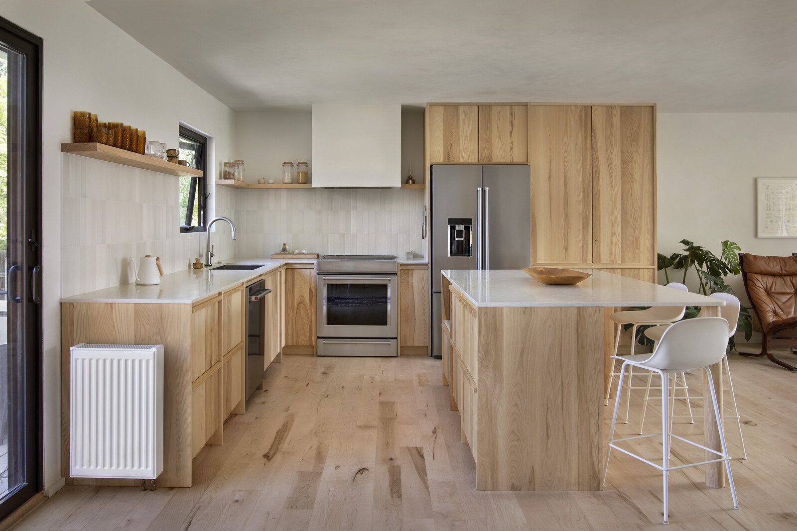 The renovated kitchen features limestone countertops, vertically oriented white Bedrosians Makoto porcelain tile, with custom ash cabinetry, maple floors, and Muuto barstools.