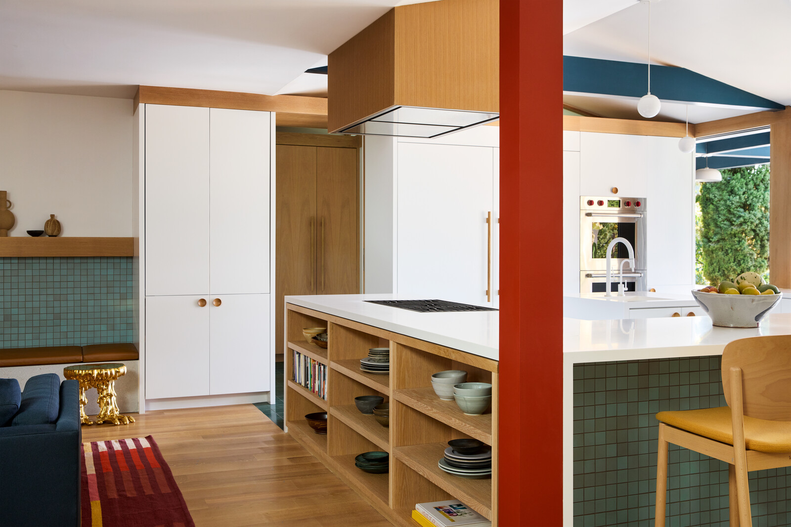 The kitchen and den exist as one continuous space, with one side of the kitchen providing added oak bookshelves for the den. A tomato-red post's color was inspired by flax plants outside.