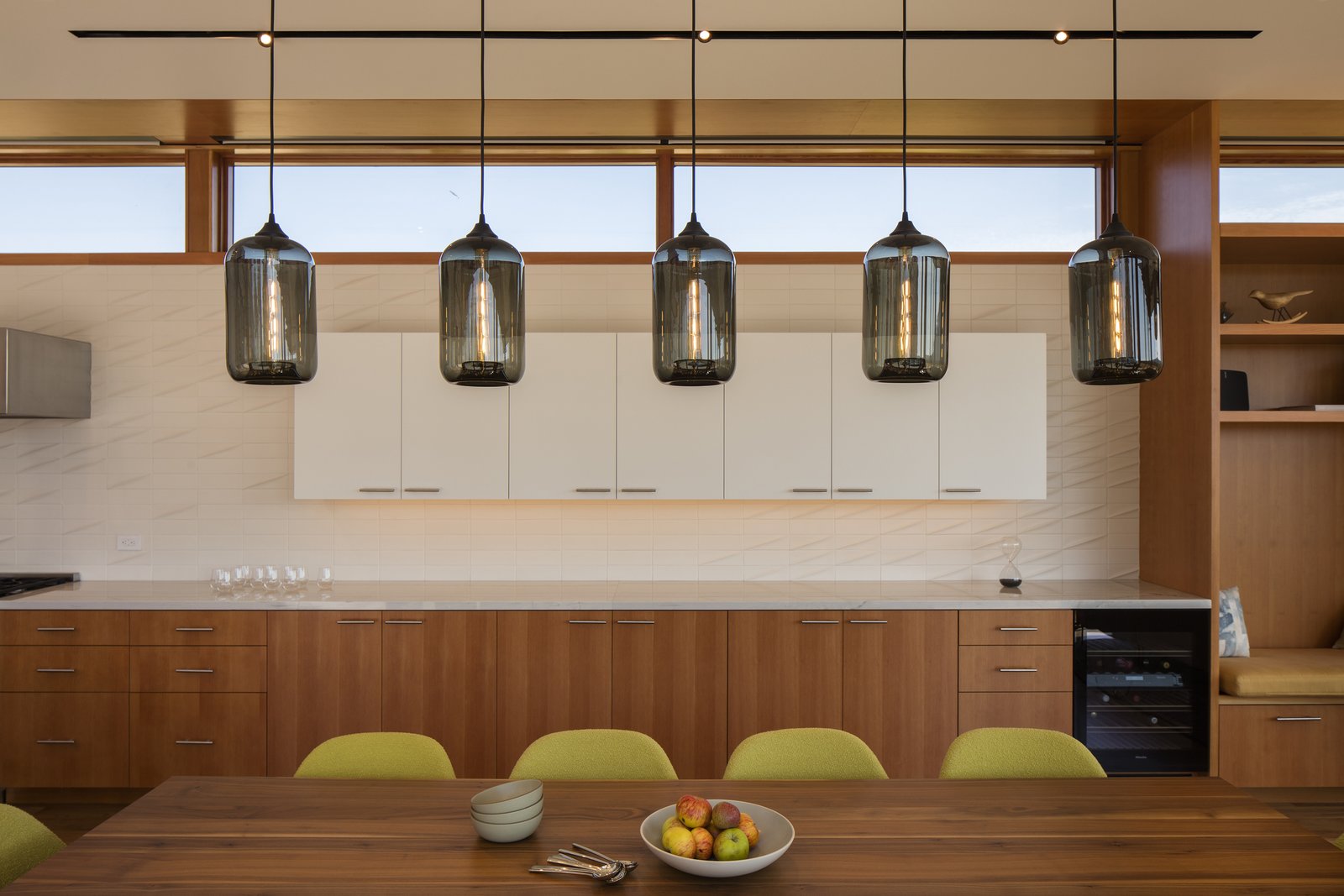 The kitchen, dining, and living room share the same space, with an open floor plan that harmoniously joins the three together. In the dining room, five Pod pendants in Gray glass hang from a Matte White Linear-5 Multi-Pendant Canopy, complementing the neutral and natural palette of beige and wood tones. The lime chairs situated around the dining table add a pop of color, inspired by the greenery just beyond the home's interior.