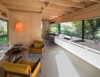 A pair of 1970s velvet-upholstered armchairs flank a midcentury-style wood table in the living area. The kitchen features a line of cabinets suspended above the epoxy resin floor. “The glazing on the east side is as long as the facade and spans eight meters,” the architect says. “It’s adjusted and harmonized with the kitchen worktop.”