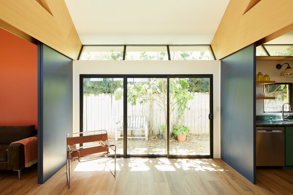The light-filled living area is flanked by a bedroom/office with an orange wall and a kitchen with green cabinetry and countertops. Tilted glass panels between the wall and the ceiling create breadth for the small home.
