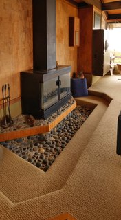 This mini conversation pit is set around a pebble-floored hearth and a steel fireplace. The warm, textured seating area reinforces the fireplace as the central feature of the room.