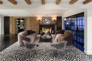 The living space features a section of ceiling with exposed wood beams, wood-paneled walls surrounding a traditional fireplace, and a wall of steel-framed windows which fill the room with natural light. 