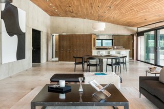 The great room comprises an open-plan living, kitchen, and dining area. The Travertine tile flooring features radiant heat. Oversized sliding glass doors lead out to an outdoor terrace, while the ceiling is paneled in teak. 