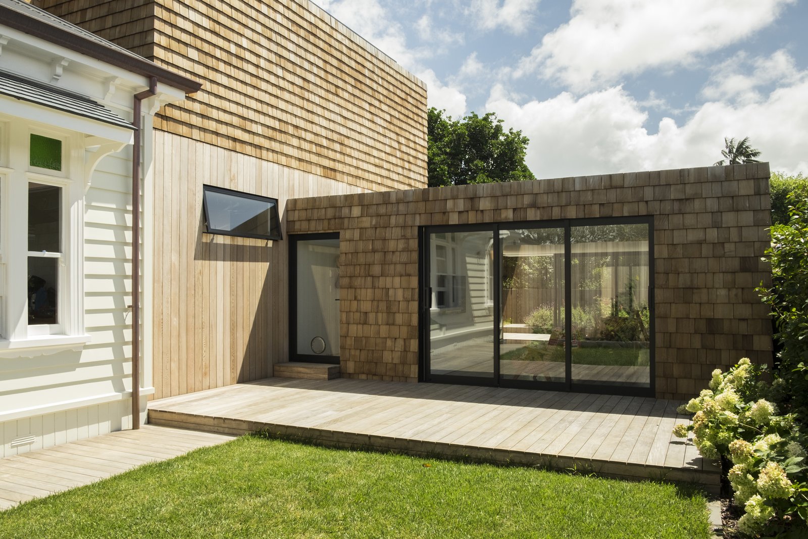 The juxtaposition of old and new architectural forms is best viewed from the courtyard between the garage and playroom. Timber shingles and vertical boarding can be seen on the sun-shading and sub-floor skirt of the villa respectively—these subordinate materials of the historic architecture are given primary roles in the new design.