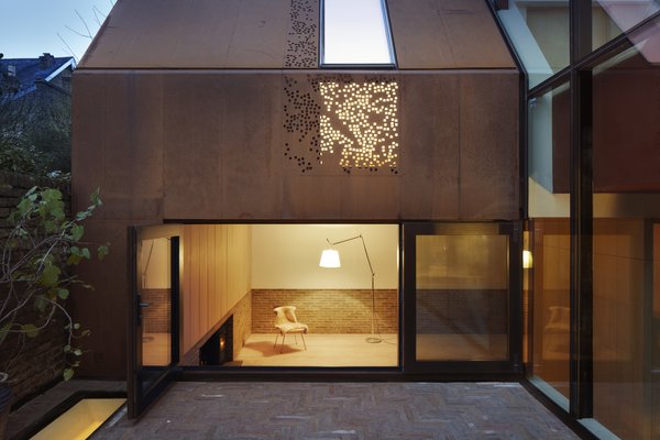 Brick and oak line the interior of the living room, where a wood-burning stove creates a welcoming ambiance. A large picture window opens onto the courtyard. Above, a perforated steel panel allows for an interplay of light and shadow.