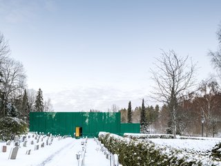 The green glass reflects the surrounding vegetation, integrating the building into the garden spaces framed by the cemetery’s hedges. 