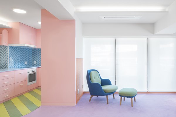 The rosy, dull pink of the kitchen cabinets bleeds into the living room but is starkly contrasted with the striped green-and-yellow floor and blue backsplash in the kitchen and furniture in the living room.