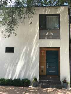 The concrete block is now covered in light-colored stucco, grey lap siding, and charcoal metal trim. The front door is painted teal and surrounded by clear-coated Maple.