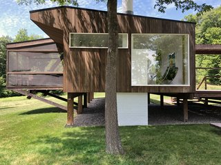 The structure appears to float above the ground. Wood stilts gently touch upon the earth, minimizing the building's footprint on the landscape. The brick chimney can be seen as it passes from the interior living spaces to the ground.