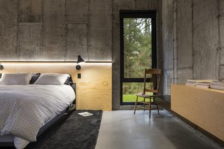 Concrete walls, a plywood ceiling, and built-in wood furnishings create sleek, minimal sleeping quarters. A plywood headboard is adorned with modern black sconces.