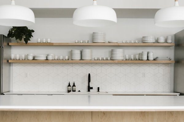 Modern white pendant lights from Schoolhouse hang above the open kitchen island. The geometric splash and open wood shelves continue into the kitchen.