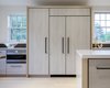 Modern home with Kitchen, White Cabinet, Wood Cabinet, Light Hardwood Floor, Laminate Cabinet, Stone Tile Backsplashe, Dishwasher, and Recessed Lighting. The cabinets are semi-custom from Brookhaven with a whitewashed face of wood-grain laminate. To customize the look, they added contrasting bleached-ash supports between each pair of cabinets to match the wood-panel hood enclosures, the cabinets above, and the floor. Photo 4 of Live-in Kitchen