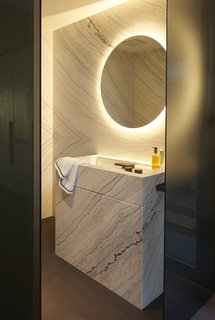 Many modern bathroom vanities use marble to elevate the style while keeping things elegant. The marble vanity seen here blends seamlessly into the marble wall behind it. Meanwhile, a striking illuminated mirror adds visual interest. 