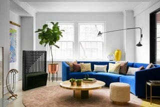 The wood herringbone floors were lightened, and bold furniture was brought in to brighten up the space. A circular custom sofa designed by MKCA, upholstered in a bright blue synthetic textile from Maharam, serves as the centerpiece of the room, readily available for family time, intimate conversation, or parties. Surrounding a vintage brass table are a sculptural chair by Charles Rennie Mackintosh and poufs upholstered in shaggy mohair.  The carpet by MKCA is made from joining two shapes of broadloom synthetic silk together.