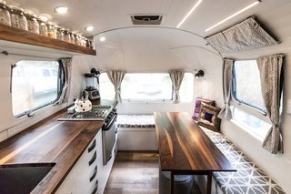 The living area is located in the front of the Airstream. It includes a full kitchen with a fridge, sink, and propane range; movable benches with integrated seating; a walnut folding table that matches the countertop; and upper shelving for dry food storage in mason jars.