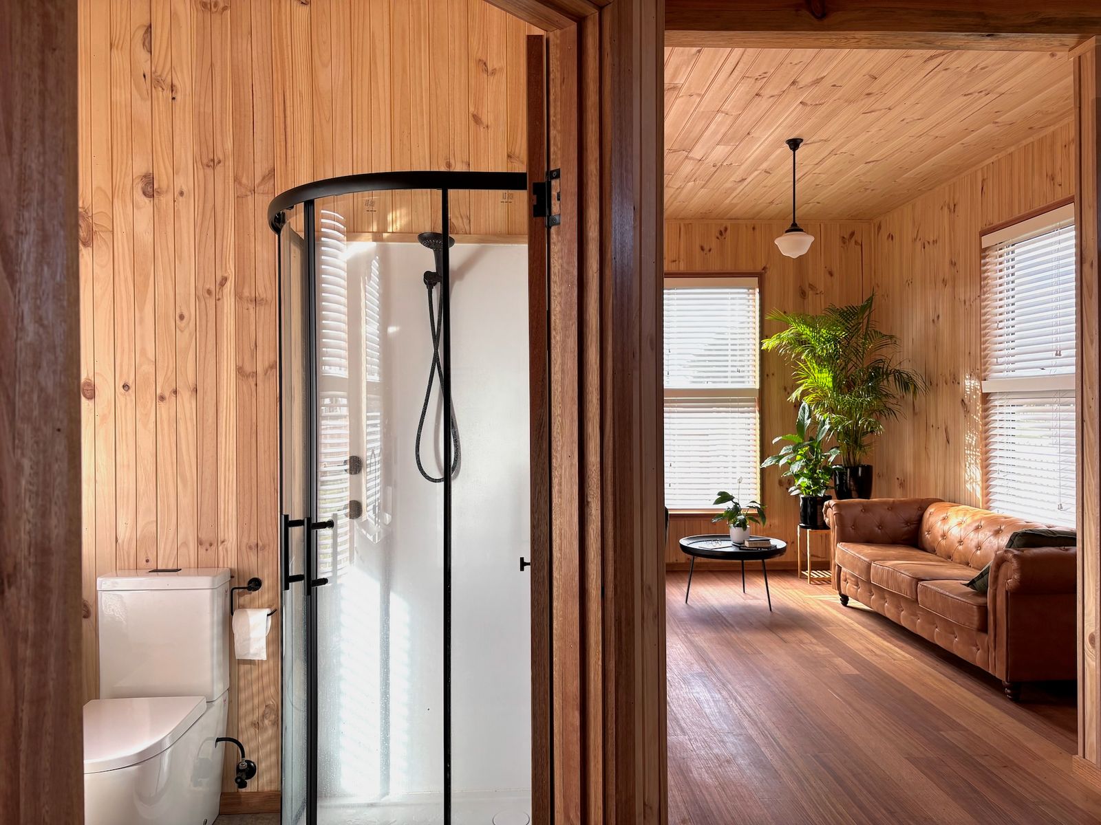 A curved glass shower enclosure and matte black fixtures give the bathroom a clean, modern finish—while the timber wall lining continues uninterrupted, echoing the simplicity and palette of the rest of the home.