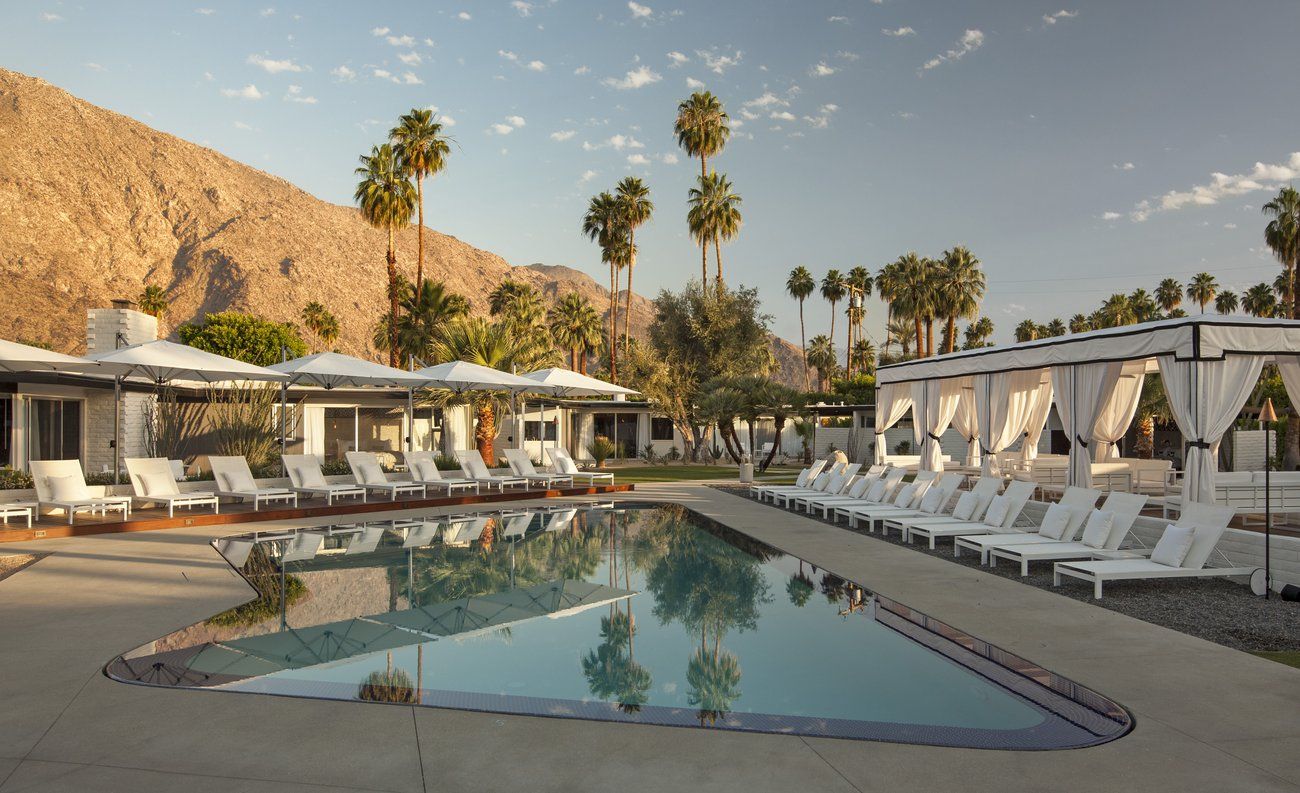 The central pool is surrounded by white loungers and private cabanas, with views of the nearby mountains and palm trees.
