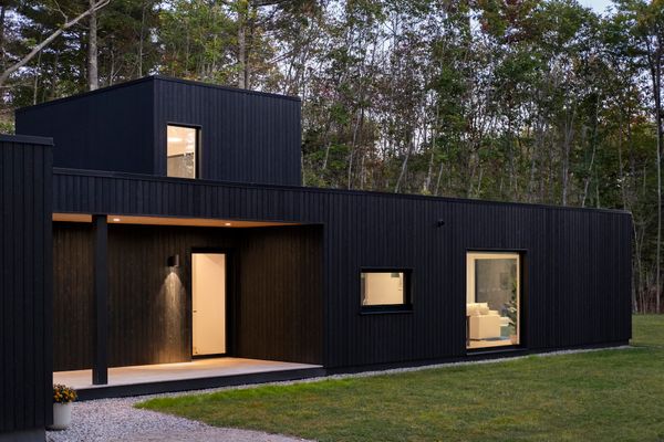 The composition of flat-roofed volumes—clad entirely in Nakamoto Forestry yakisugi sourced from PEFC-certified, air-dried Japanese cedar—reads as a single dark form against the forested landscape, punctuated by carefully placed openings that offer glimpses of the bright interior. At twilight, the entry facade (as seen here) becomes illuminated by warm light spilling from the covered porch and windows onto the gravel path.