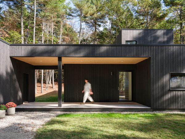 A generous covered entry porch provides a sheltered connection between the main house and a small garage and workshop. The warm-toned wood soffit creates a striking contrast against the charred Japanese cedar cladding, while the open breezeway beyond frames a view through to the woods.