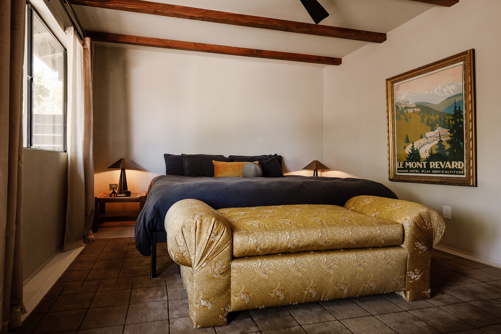 The updated primary has exposed wood beams and an eclectic mix of furniture and art. The bed frame is from Tuft and Needle, and the bench is vintage that Kelly reupholstered in a gold floral pattern.