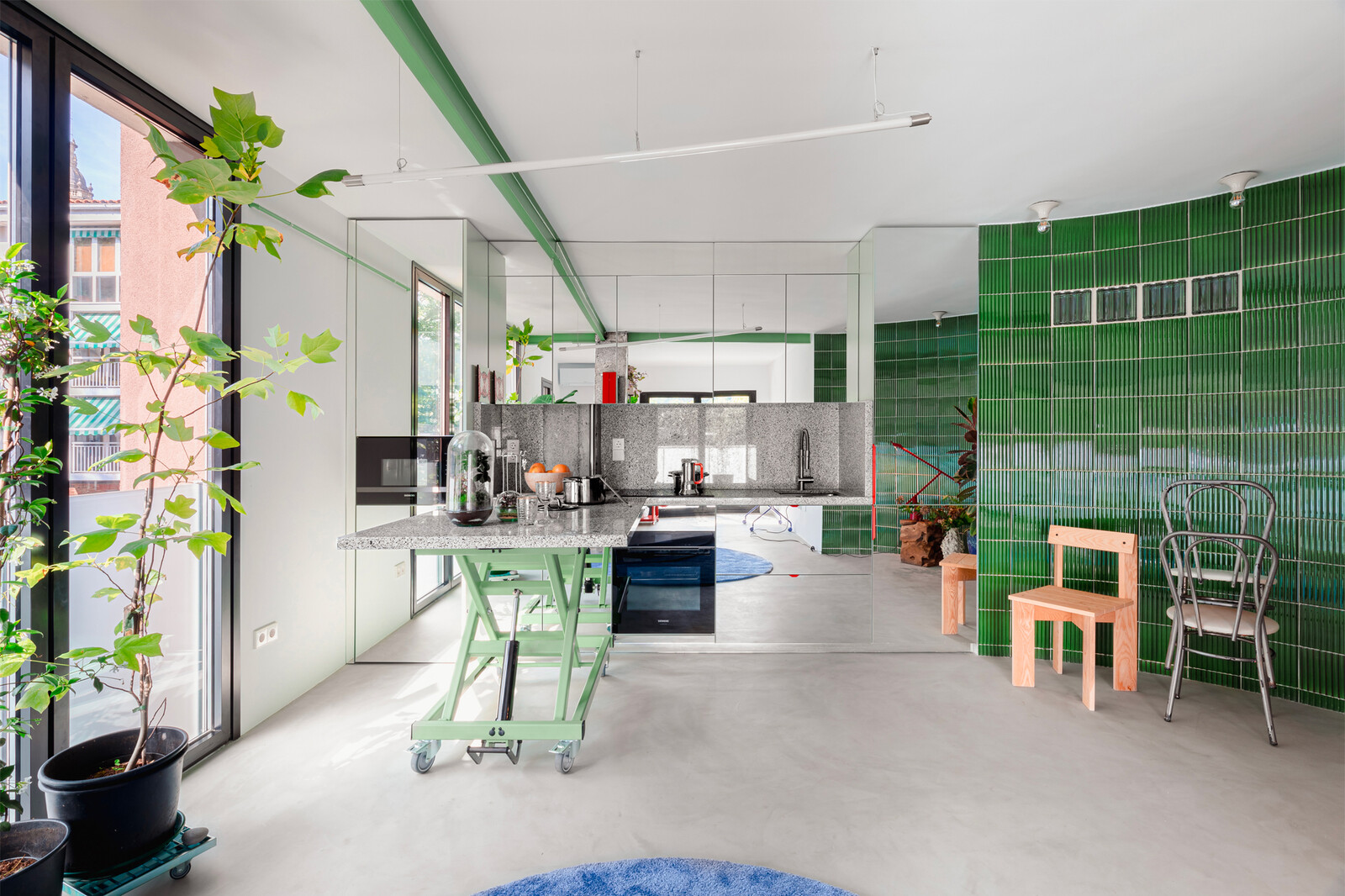 The kitchen is tucked into a corner off the curved green wall with mirror-clad cabinetry, helping it almost disappear into the space. The island is on wheels and incorporates a hydraulic piston so that it can be moved and raised and lowered for different uses in the space.