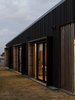 Modern home with Exterior, Butterfly RoofLine, Beach House Building Type, Wood Siding Material, Hipped RoofLine, House Building Type, and Metal Roof Material. Glowing at dusk, the large glazed sliders are bordered with black steel hoods that aid in shading with interior lined natural wood that contrasts with the dark battened ply cladding. Photo 8 of The Crest House