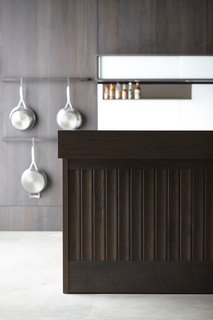 The test kitchen is outfitted with their Opencase Wall System, configured with two pot racks and a sliding glass wall unit that includes integrated LED lighting. The Backsplash System is constructed of solid white acrylic and features a stainless steel shelf. Finally, the gray walnut island is finished with an impeccable tongue-and-groove paneling, a recent addition to Henrybuilt’s growing range of products.