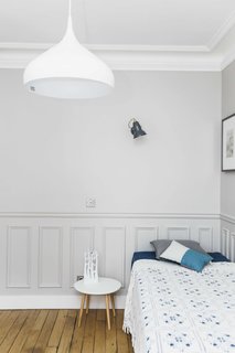 The charm of original wood floors, wall paneling, and thick crown molding is brought into the 21st century with a coat of crisp white paint and soft gray walls. Minimalist furnishings and fixtures, like an undulating white pendant and a small white bedside table, keep it feeling modern.