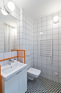 The bathroom features graphic eye-catching Vives floor tiles surrounded by Opoczno wall tiles and clean white plumbing fixtures. The custom-designed orange sink frame adds a whimsical pop of color.