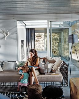 iT House, Joshua Tree - Photo 7 of 9 - Taalman sits with their daughter, Oleana, in the living room on a metal lounge designed by Kenneth Cobonpue. Glass walls emphasize views and greatly expand the sense of space in the 1,100-square-foot house. Through the glass wall behind her is the fire court and behind that the master bedroom.