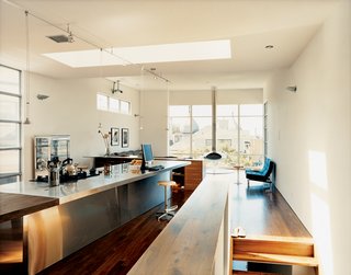 A long, narrow rectangle functions as a kitchen, dining, working, and general gathering area. Skylights set at a 90-degree angle to the workstation boost the dynamism in the room and cast a bright glow over the unit's walnut-and-steel seam.