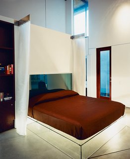 The glass platform bed is backed by a headboard of two sheets of lucite embedded with aquamarine wool from Maharam. Hanging pillow-side, electroluminescent draperies glow in the dark.