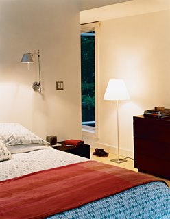The master bedroom has a number of windows in varying shapes and sizes, providing unique views to the woods outside. The bed, chest of drawers, side tables, and geometric linens are all from West Elm. The Tolomeo wall lamps are by Artemide.