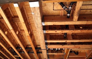 The ceiling in the living room shows the network of plumbing. Small copper pipes carry water and the black cast iron pipes carry waste. Plumbers and electricians drill through freshly installed studs and beams as they find a route for their pipes, conduits, and wires. Engineering calculations are used to locate areas where holes through the beams will have minimal structural impact.