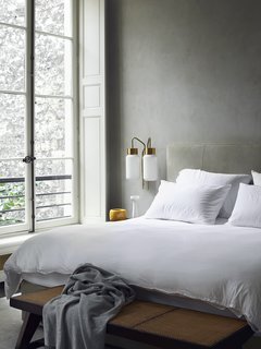 Architect Joseph Dirand's bedroom boasts sandy gray walls and upholstered headboard. The white bed linens help reflect daylight into the space and the Azucena sconces offer task illumination.